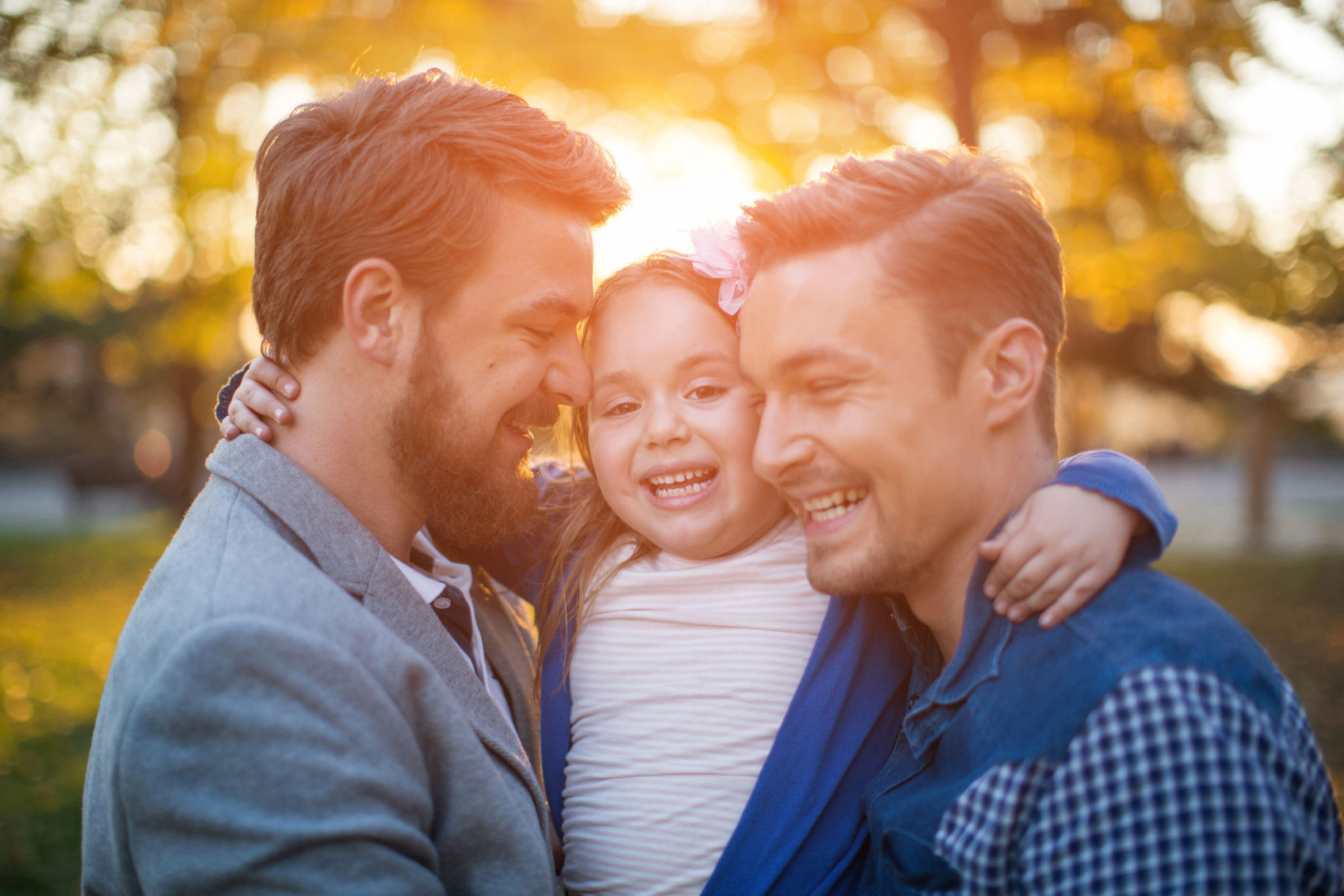 A photo of two proud fathers holding their happy child with the sunset in the background.